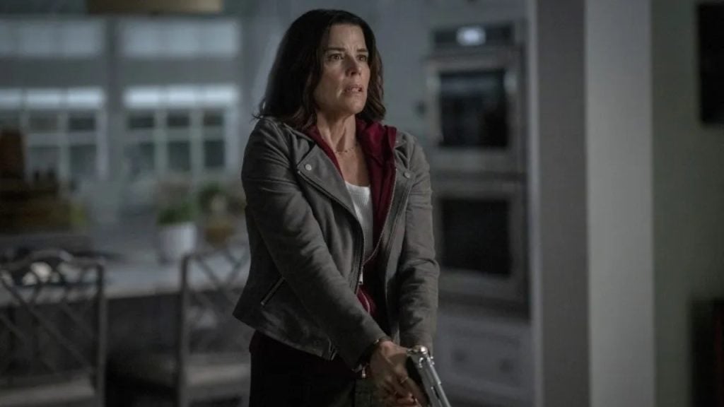 A woman with dark hair and a gray jacket stands indoors, holding a handgun with both hands, looking tense and alert.