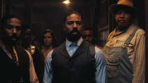 A group of people, dressed in early 20th century attire, stand together indoors with serious expressions under dim lighting.