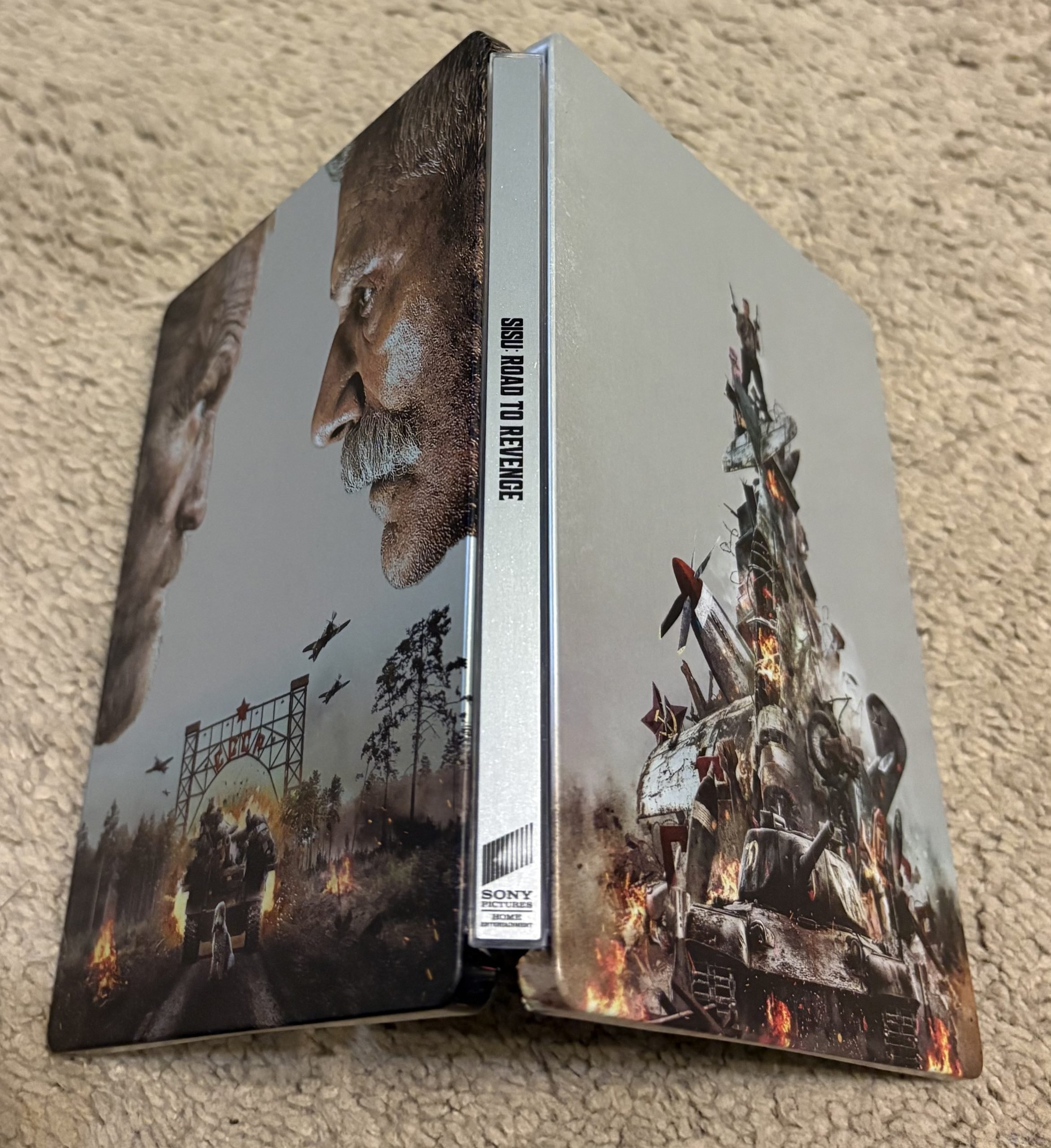 A steelbook case for "Sisu: Road to Revenge" shows two men facing each other on the front and an explosive war scene with a tank and missiles on the back.