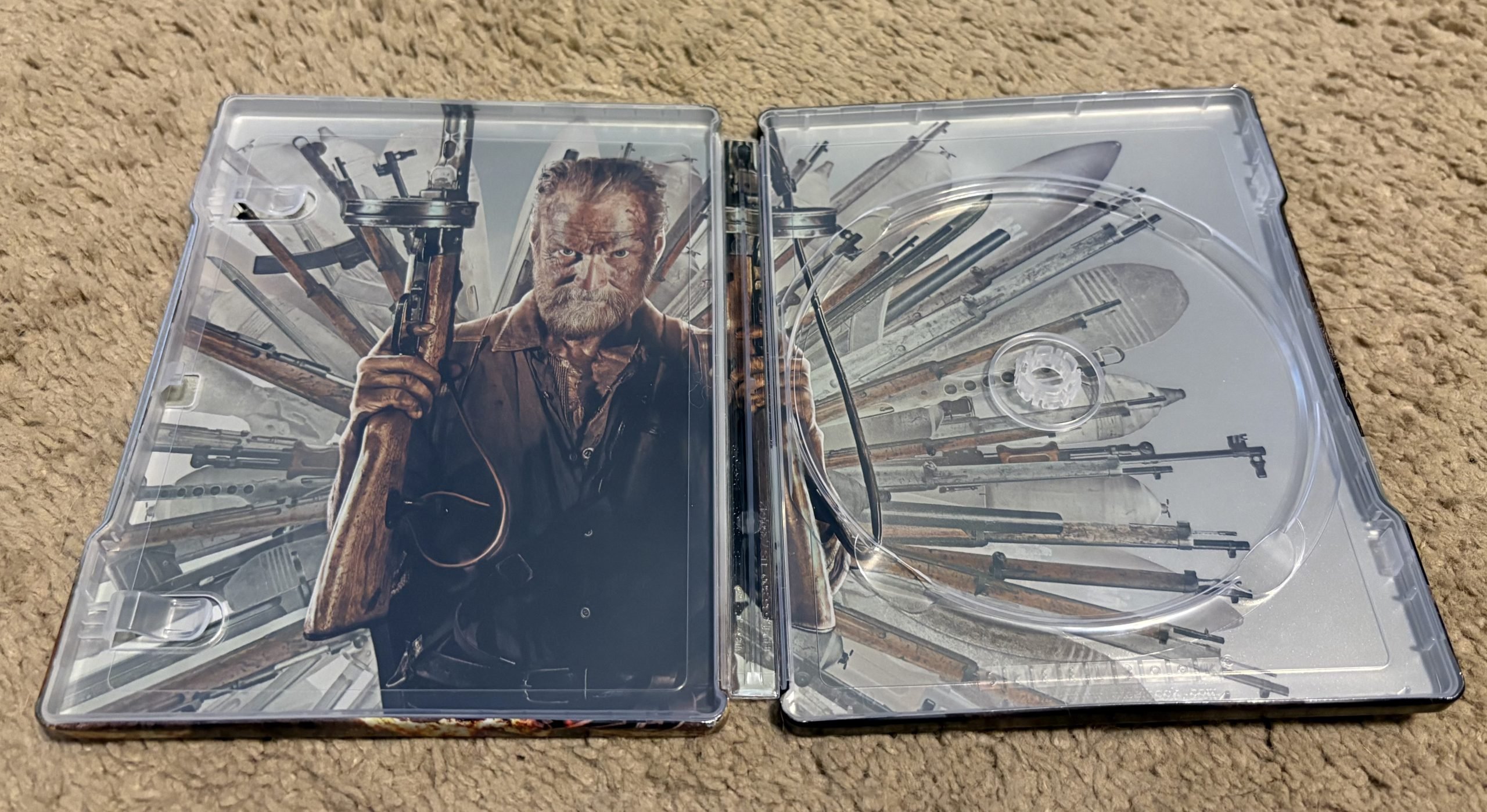 Open Blu-ray case featuring artwork of an older man holding a gun, surrounded by various rifles and firearms, on a beige carpeted surface.
