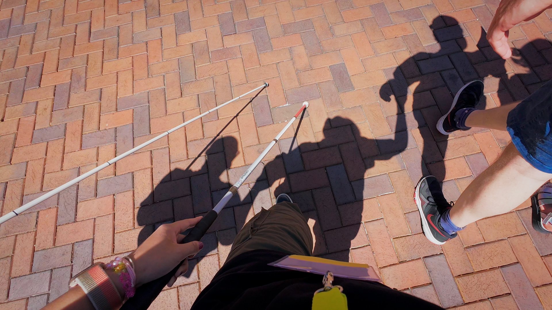 A person holding a white cane walks on a brick path, with their shadow and the shadow of another person visible on the ground.