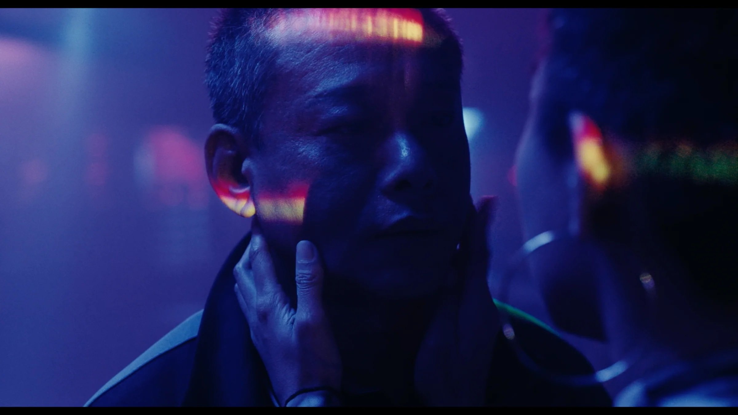A person holds another person's face in their hands under blue and purple lighting with colored reflections across the man's face.