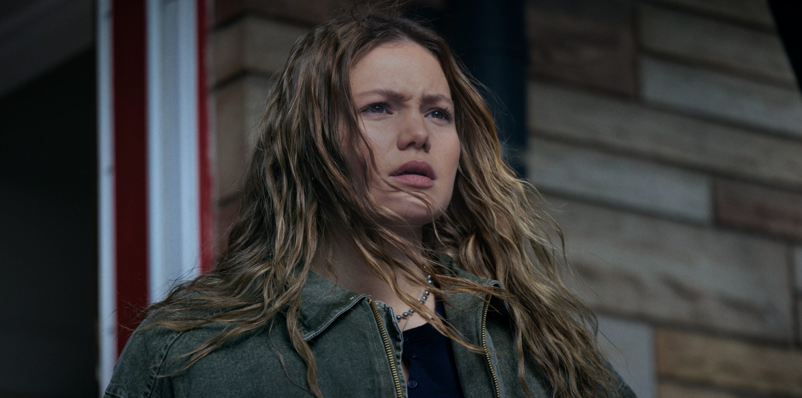 A woman with long, wavy hair wearing a green jacket stands outside in front of a brick wall, looking ahead with a concerned expression.