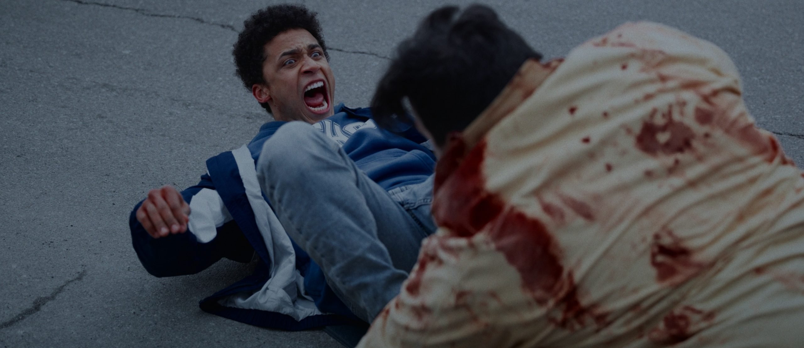 A young man in a blue jacket lies on the ground looking frightened while another person in a bloodstained shirt crouches nearby on a concrete surface.