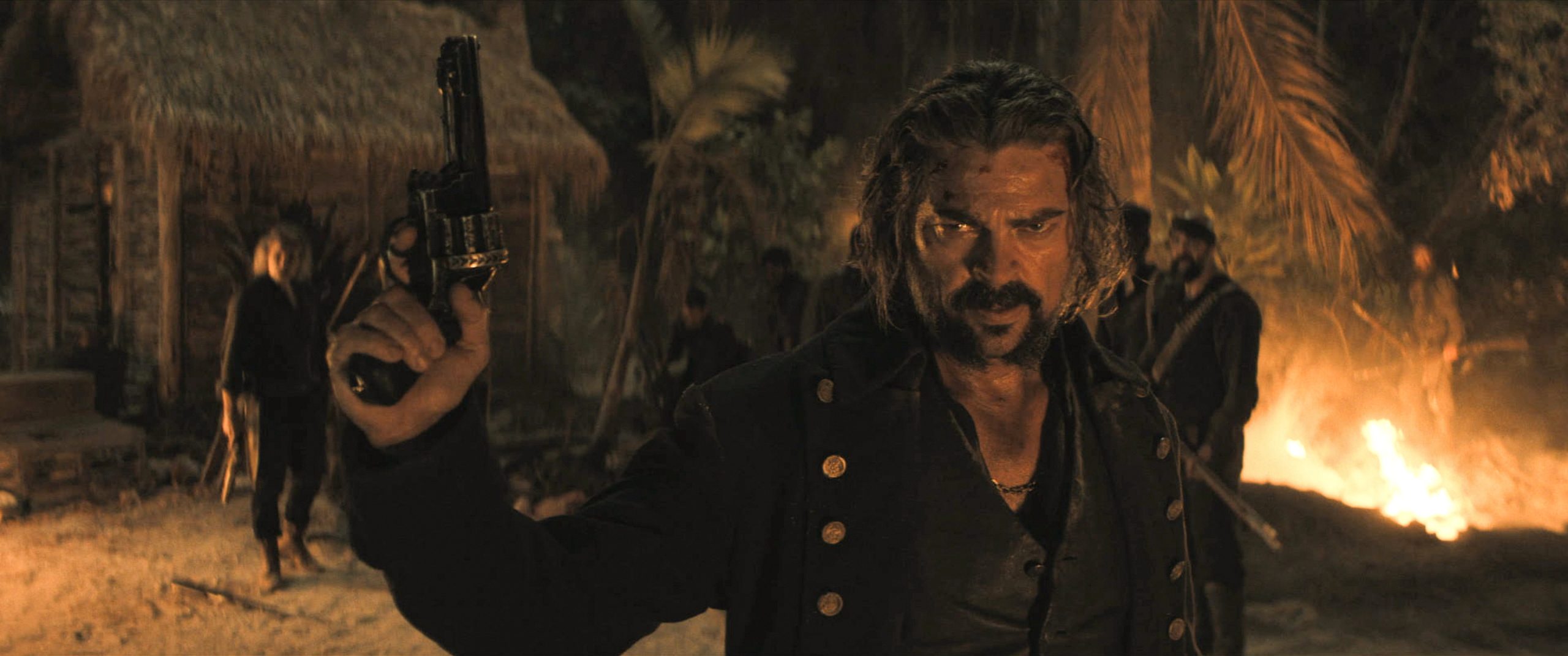 A man with long hair and a mustache holds up a revolver in an outdoor scene at night, with fire and palm trees in the background.