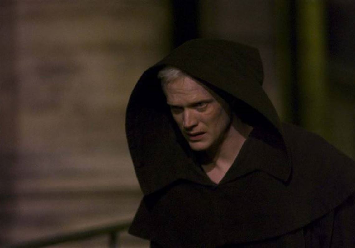 A person with pale skin and white hair wears a dark hooded cloak, walking outdoors at night with a serious expression.
