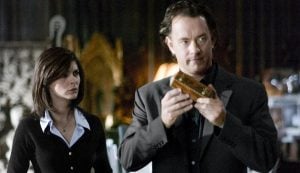 A man in a suit examines a rectangular artifact while a woman in a black sweater stands beside him, both appearing focused in a dimly lit room.