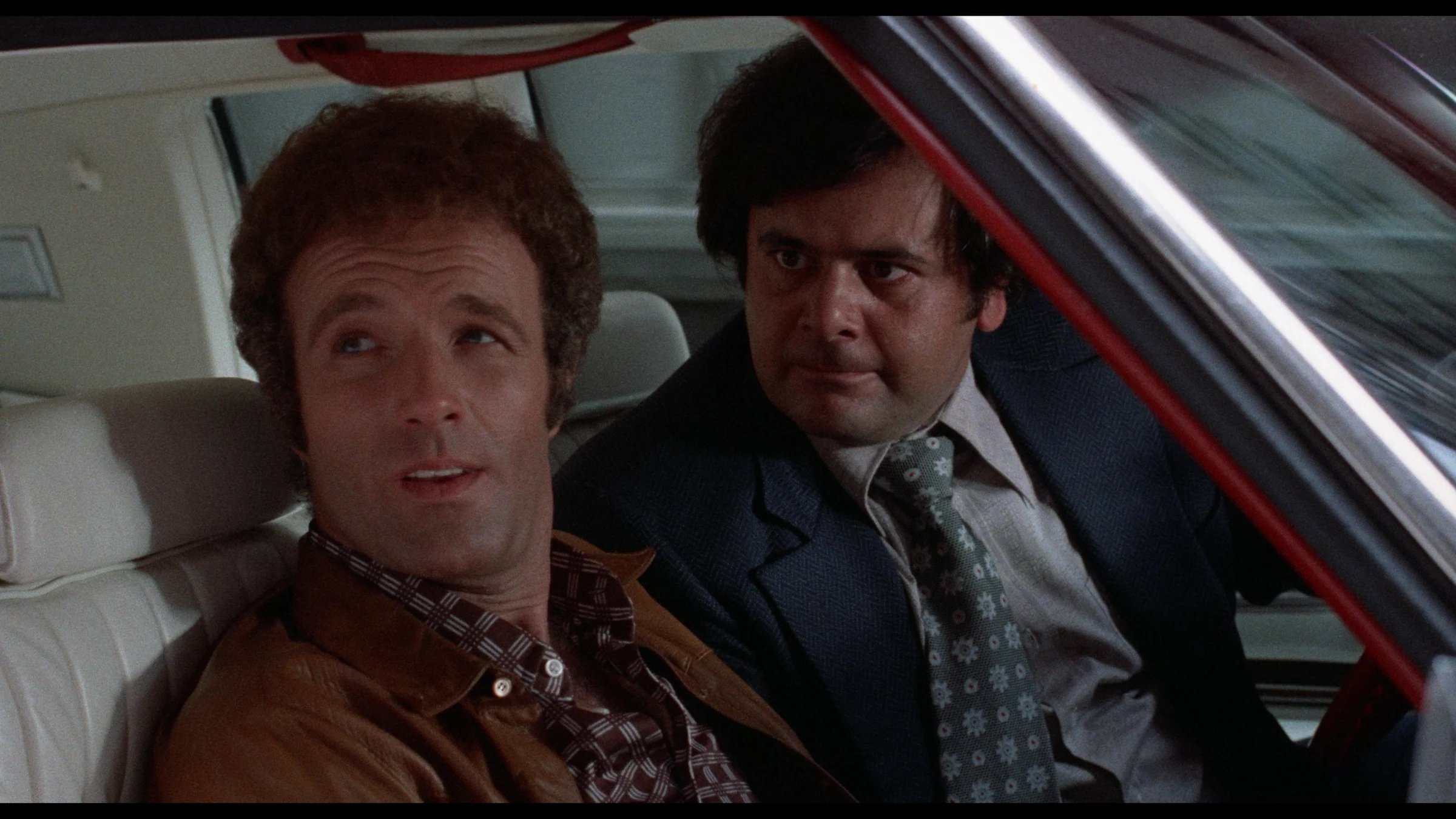 Two men sit in the front seat of a car; the man on the left looks out the window while the man on the right leans in, appearing to listen or speak.