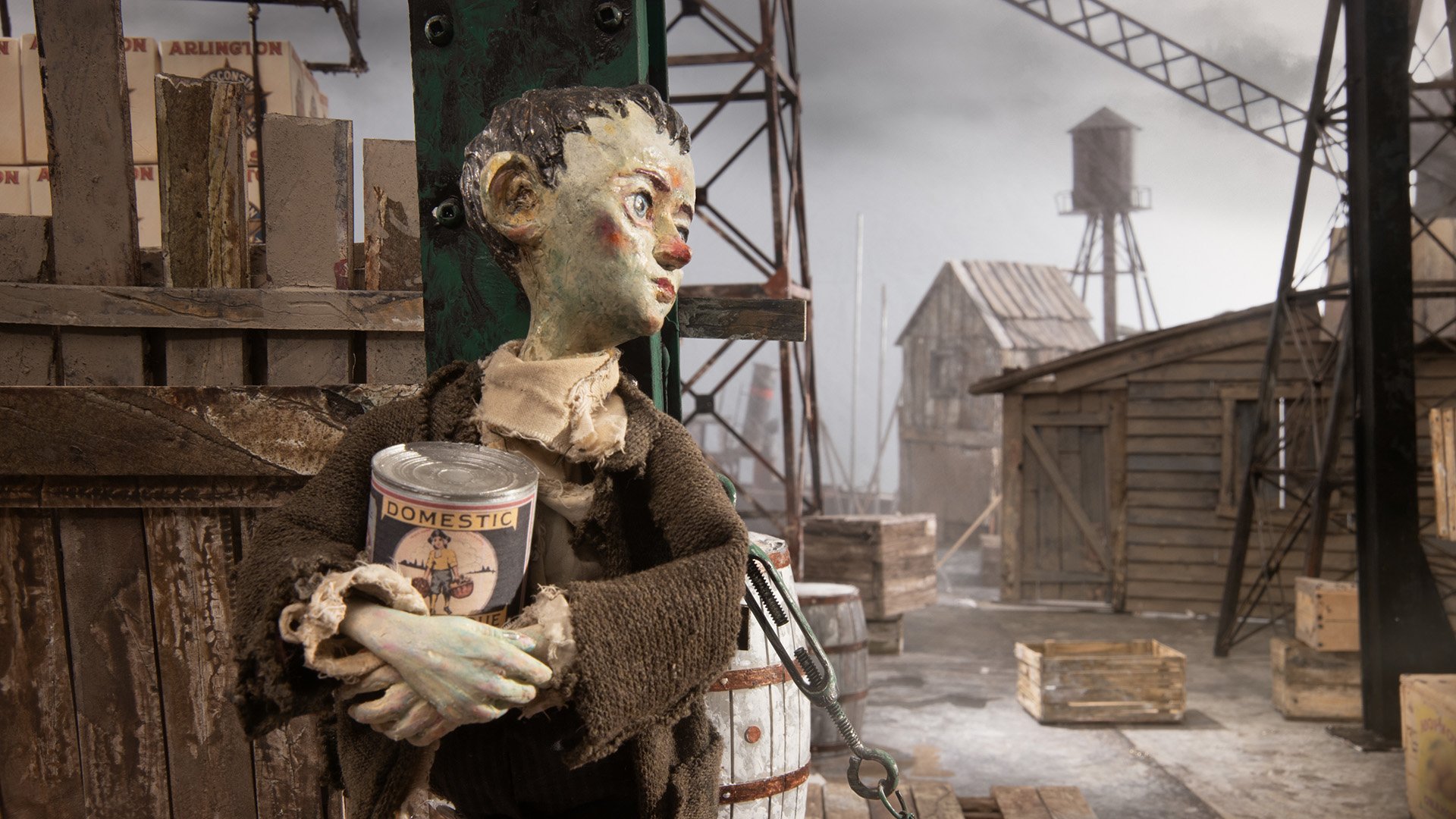 A stop-motion puppet figure in tattered clothing holds a can labeled "Domestic" while standing in a rundown, industrial dockyard setting with wooden crates and buildings.