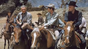 A group of men wearing cowboy hats and western attire sit on horseback outdoors, with mountains and dry trees in the background.