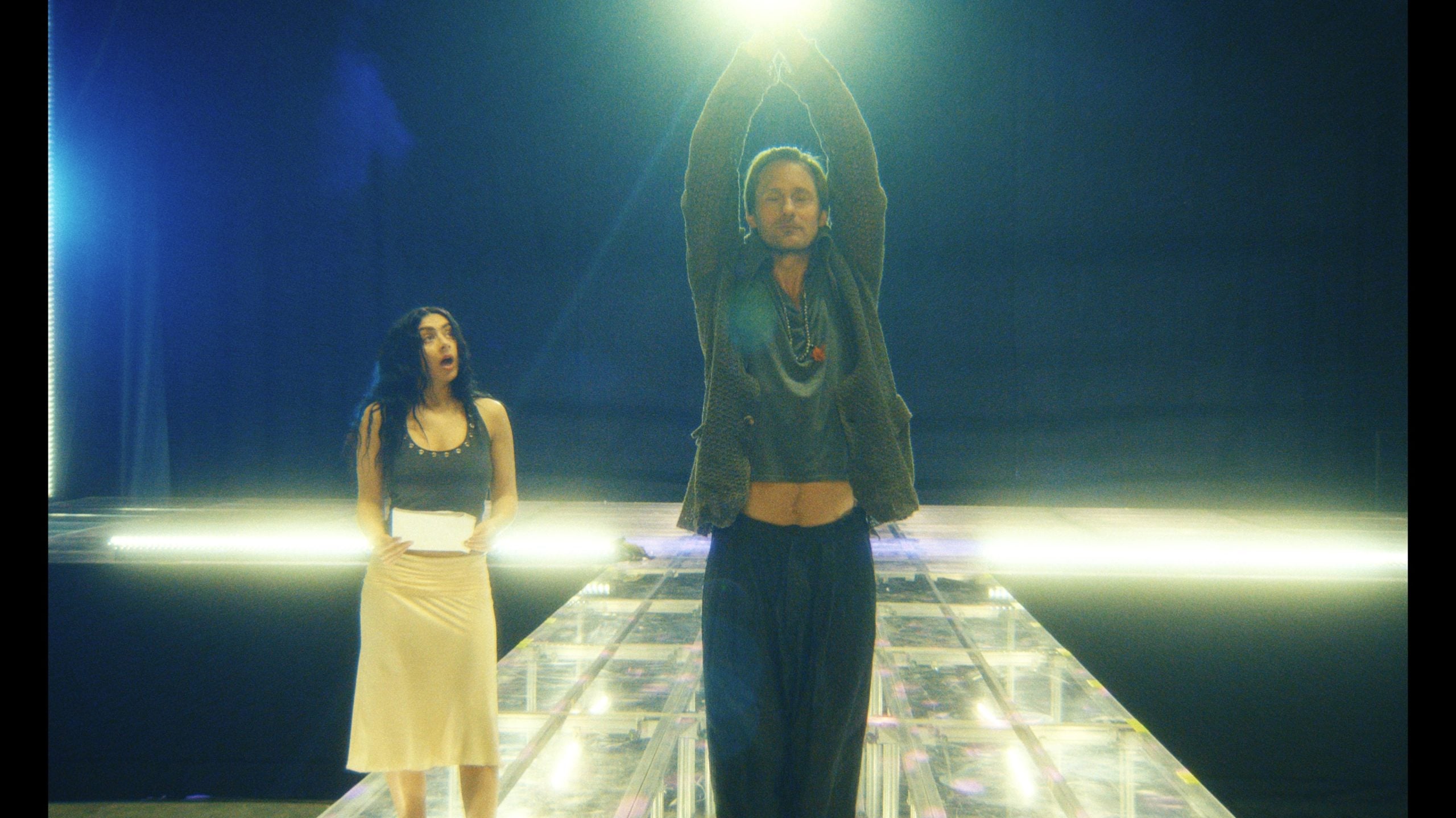 A woman stands holding a notepad while a man stands on a lit platform with arms raised, facing forward, under bright stage lights.