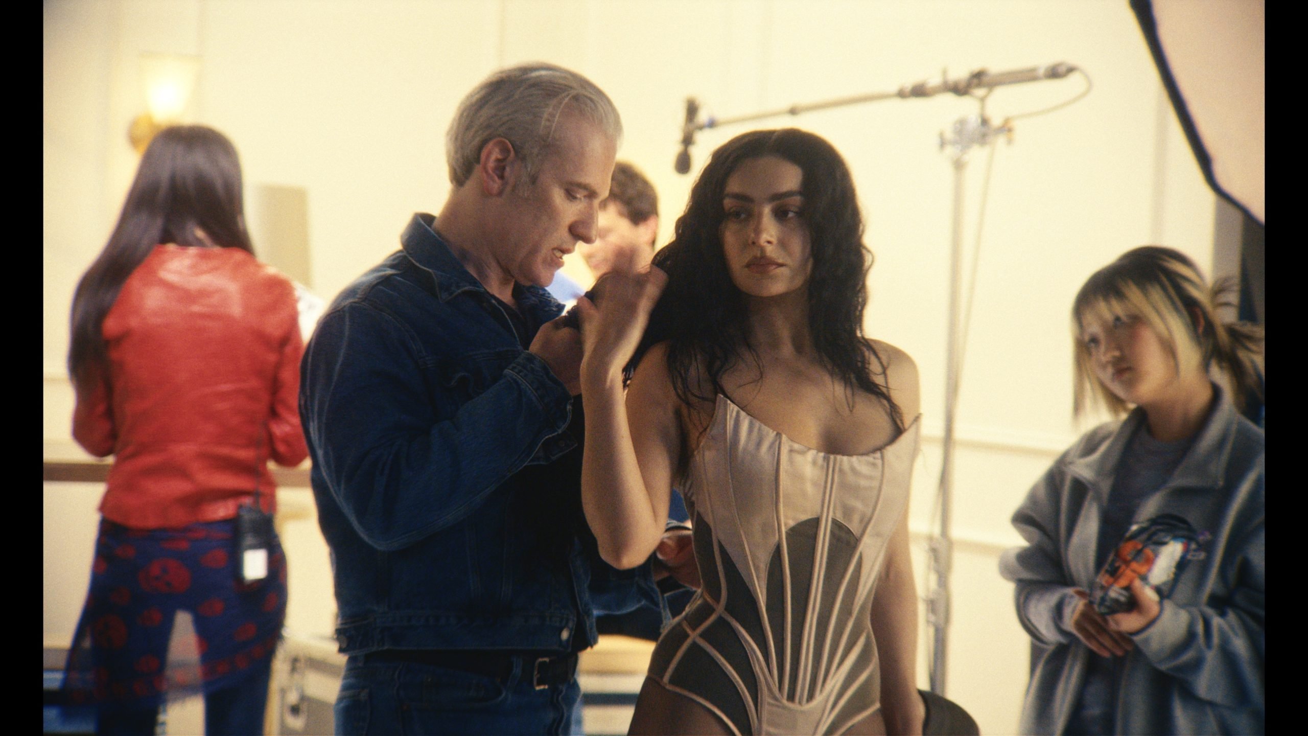 A woman in a corset stands as a man adjusts her hair. Two other women are in the background, one holding a camera. Studio lighting and equipment are visible.