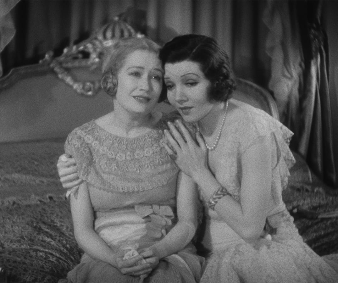 Two women in vintage dresses sit close together on a bed, one comforting the other. The scene appears to be from an old black-and-white film.
