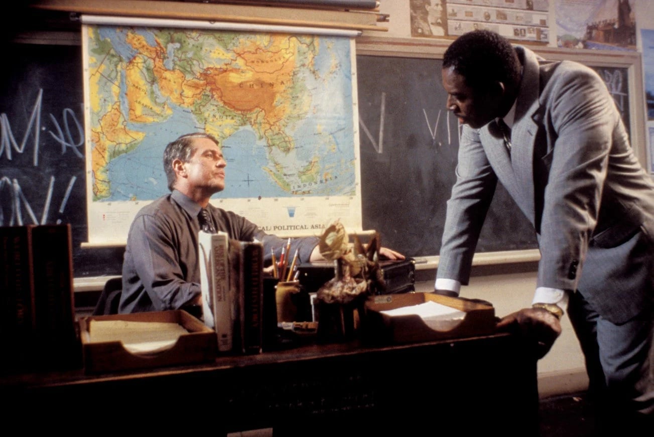 Two men in a classroom talk; one sits at a desk with books and papers, while the other stands, leaning forward. A world map and chalkboard are visible in the background.