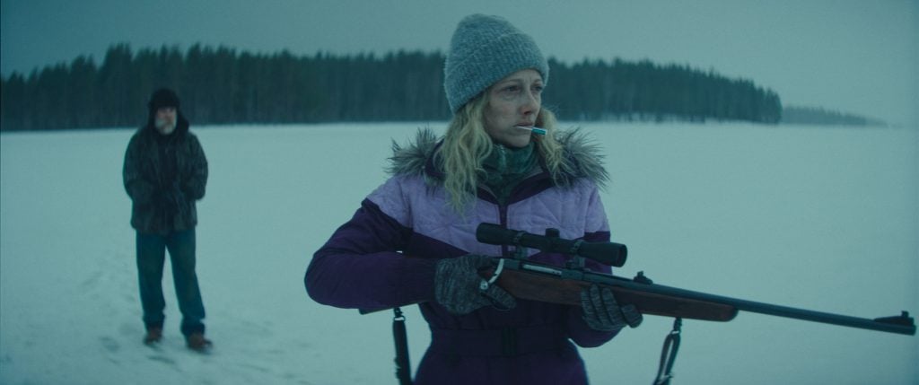 A woman in winter clothing holds a rifle and smokes a cigarette on a snowy landscape, while a man stands in the background.