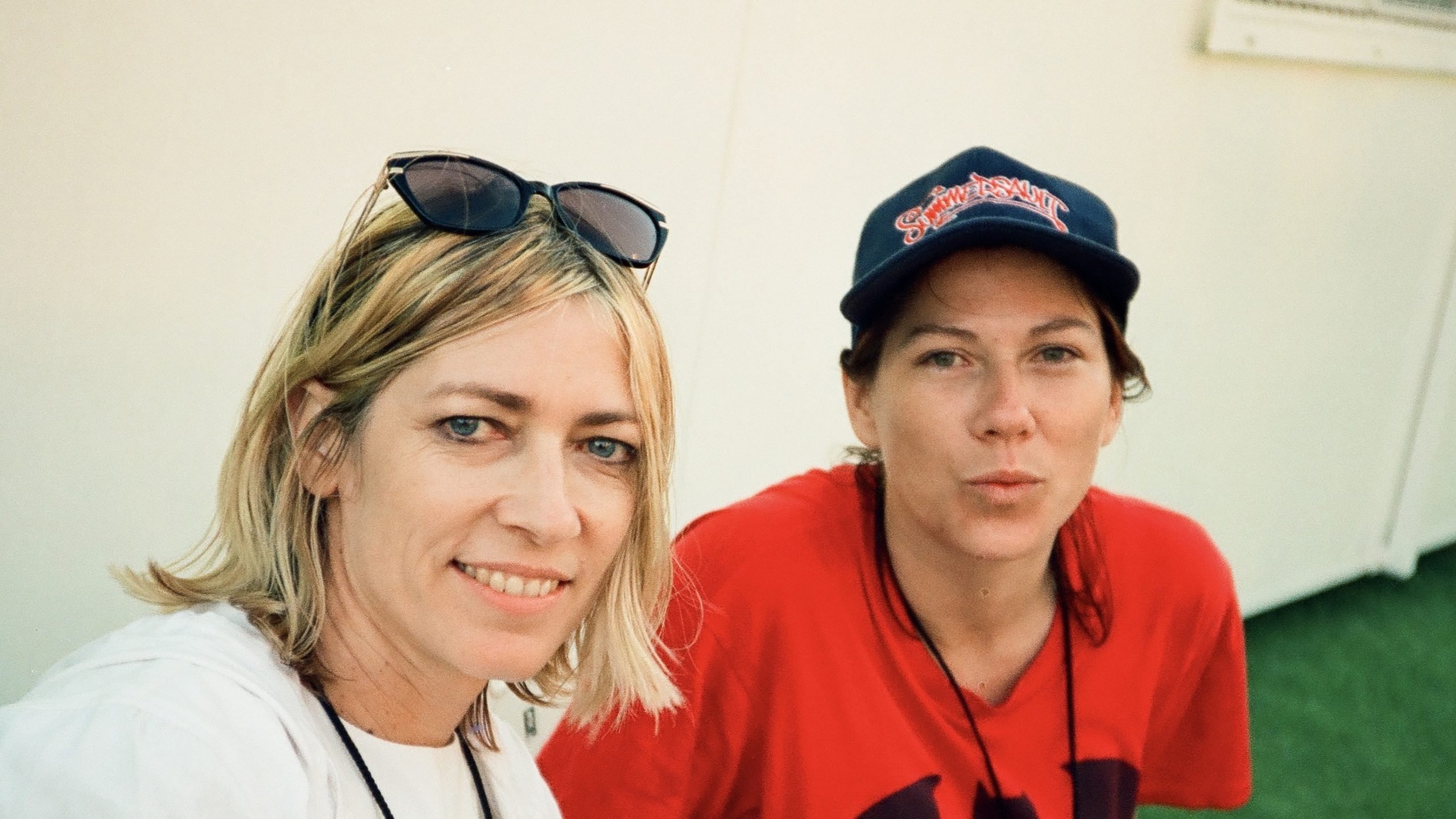 Two women are posing for a photo; one is smiling and wearing sunglasses on her head, the other is wearing a navy cap and red shirt, both in front of a plain light-colored background.