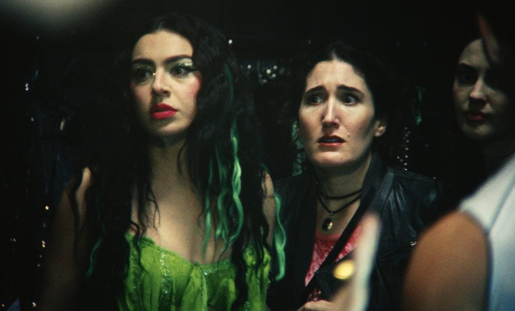 Two women stand indoors; one wears a bright green outfit with green streaks in her hair, while the other looks concerned and wears a black jacket.