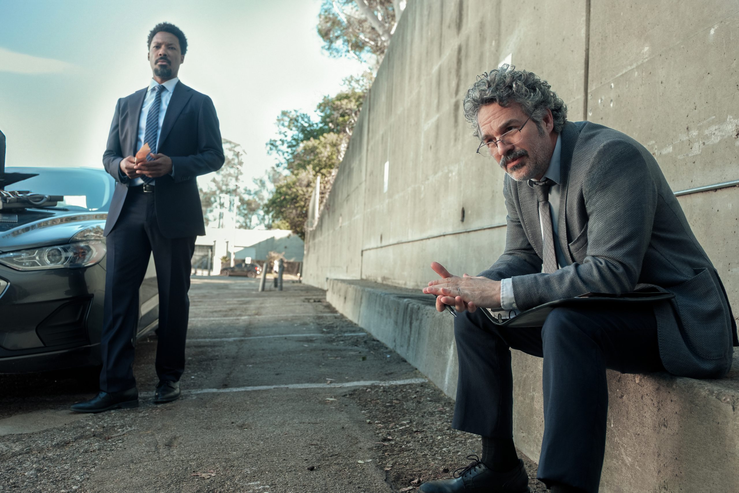 Two men in suits, one standing near a car and the other sitting on a concrete ledge by a wall, appear to be having a serious discussion outdoors.