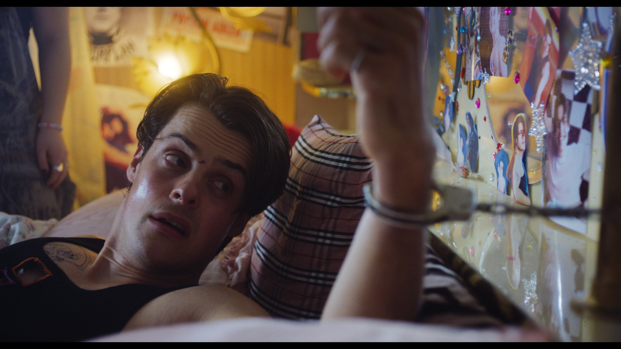 A young man in a black tank top lies on a plaid pillow, looking up at a collage-covered wall adorned with photos and lights.