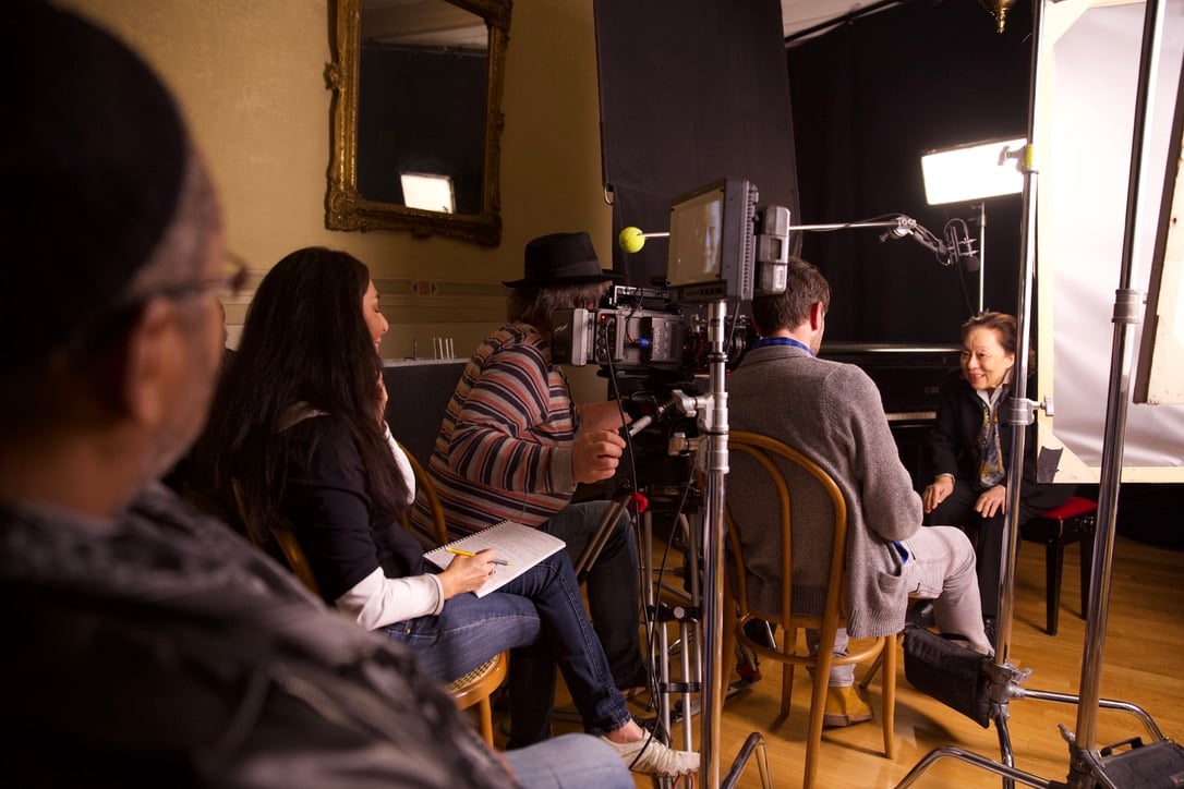 Four people are filming an interview in a studio with professional lighting and camera equipment; one person is speaking while others observe or take notes.
