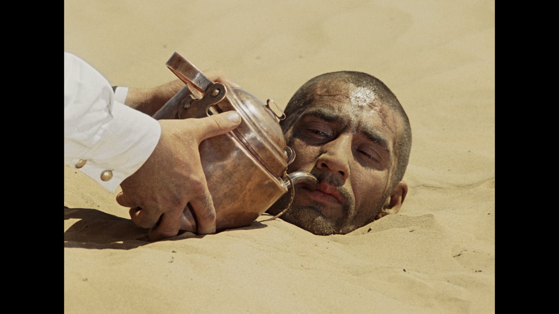 A man’s head is buried in sand while another person holds a metal teapot near his mouth.