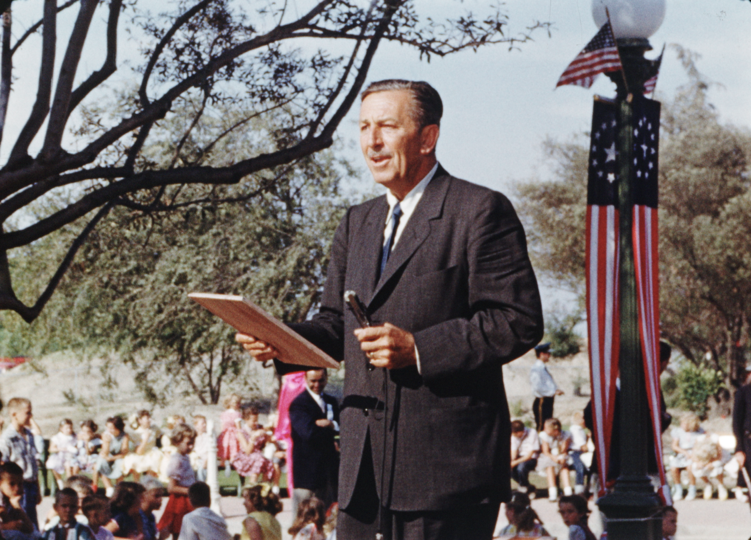 A man in a suit stands outdoors holding a plaque and microphone, with a crowd seated behind him and American flags on a nearby lamppost.