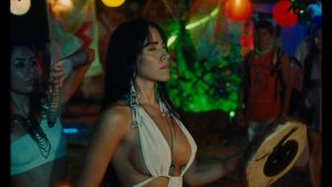 A woman in a white halter top holds a circular object, standing among colorful lights and decorations at an outdoor nighttime event with other people present.