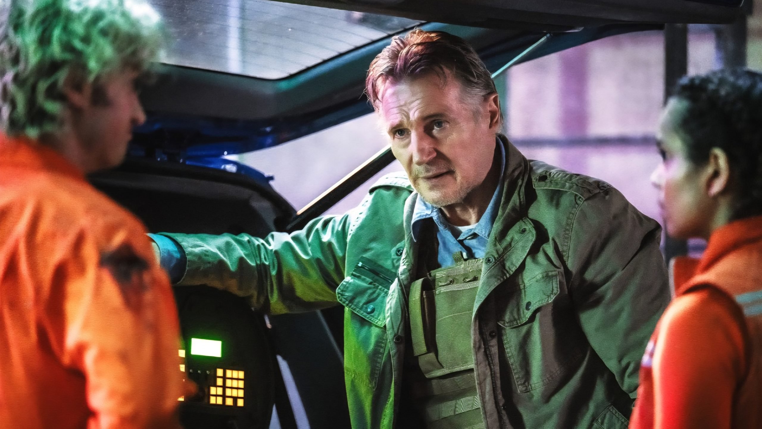 A man in a green jacket and tactical vest talks to two people in orange jumpsuits beside a vehicle with visible machinery.