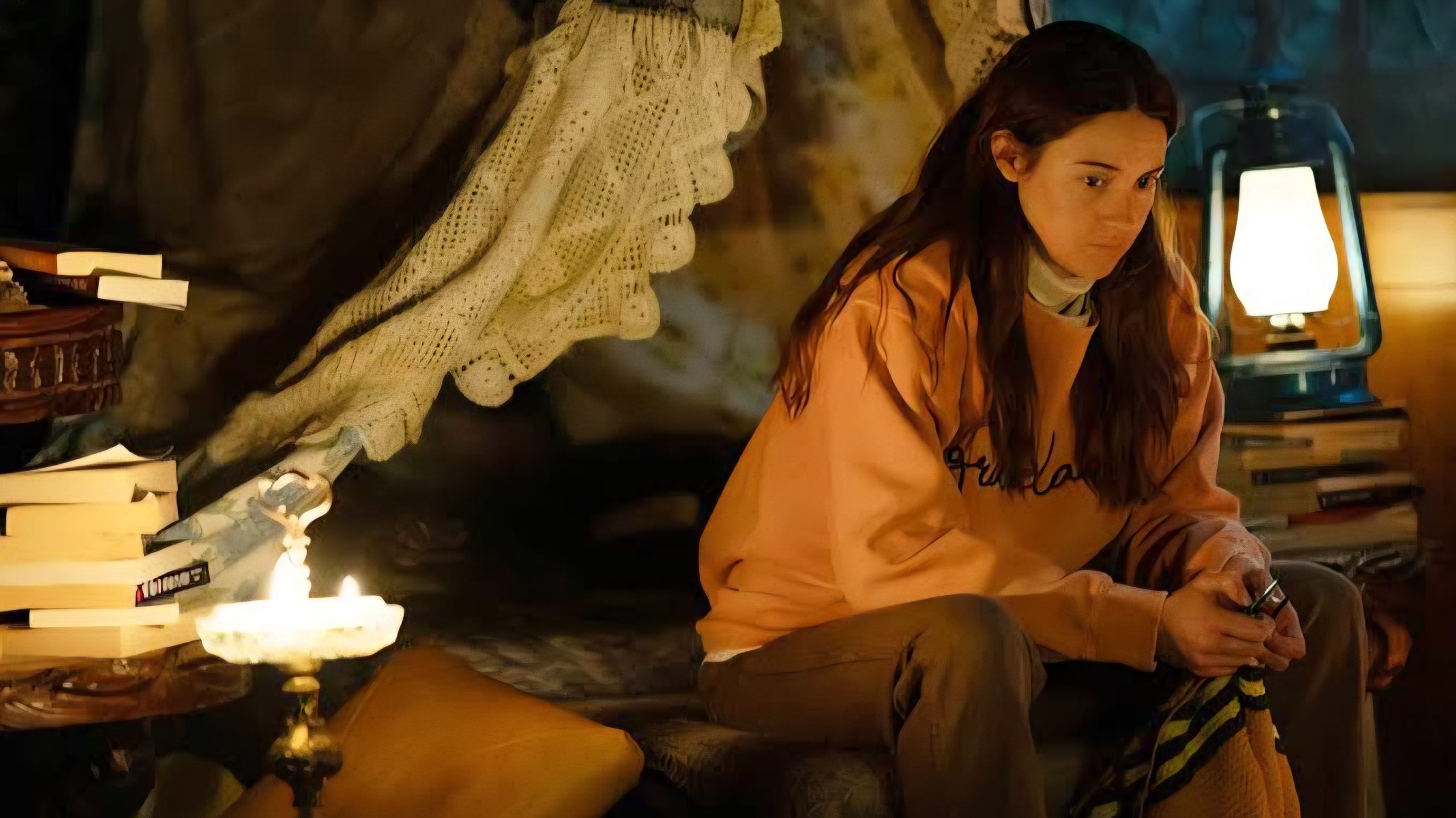 A woman in a peach sweatshirt sits indoors by a lit oil lamp, surrounded by books and blankets, looking thoughtful.