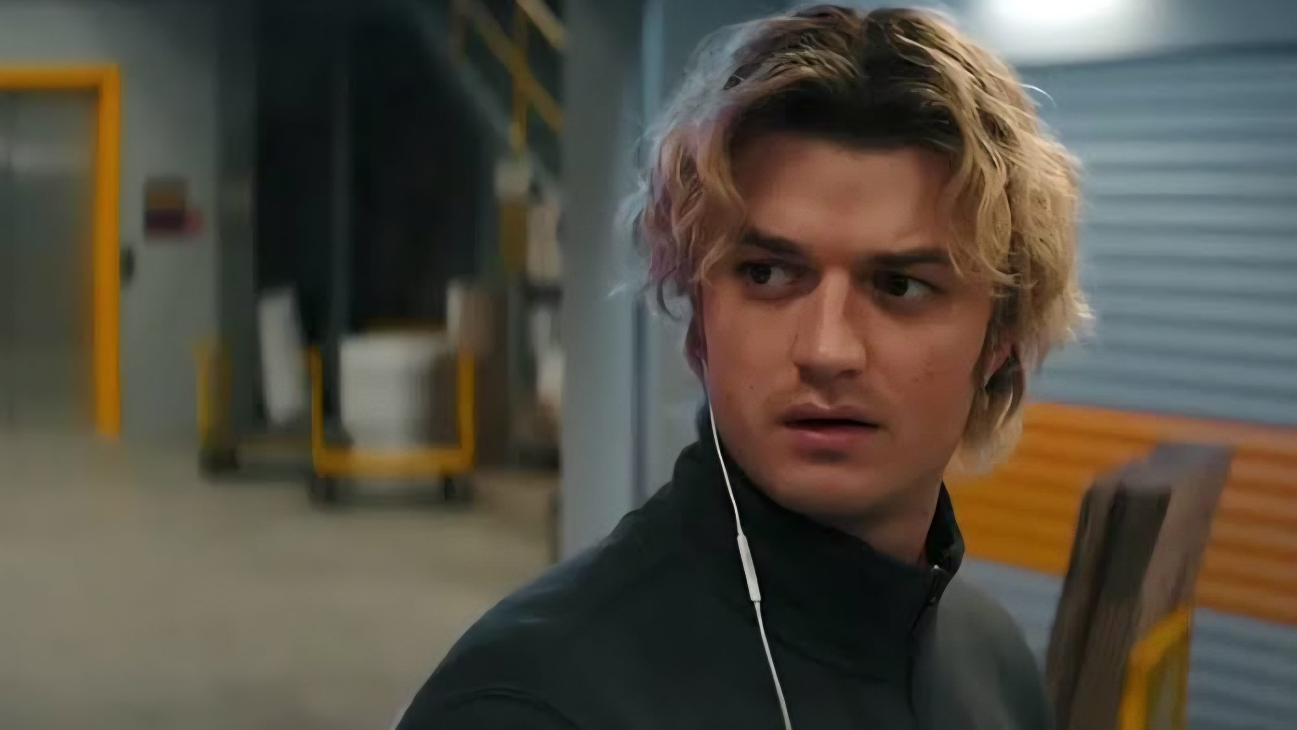 A young man with wavy blond hair and earbuds looks to his left in an industrial setting with blurred background details.