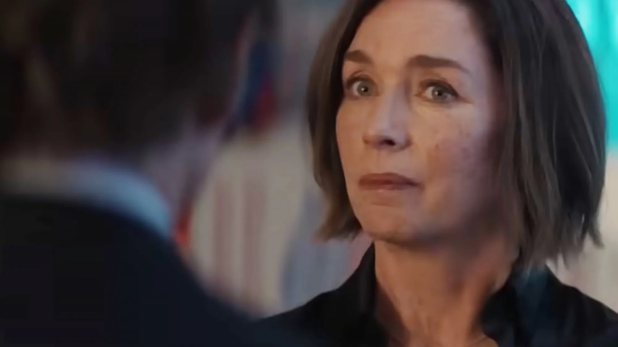 A woman with short brown hair and a serious expression looks at a person in the foreground, who is facing away from the camera.