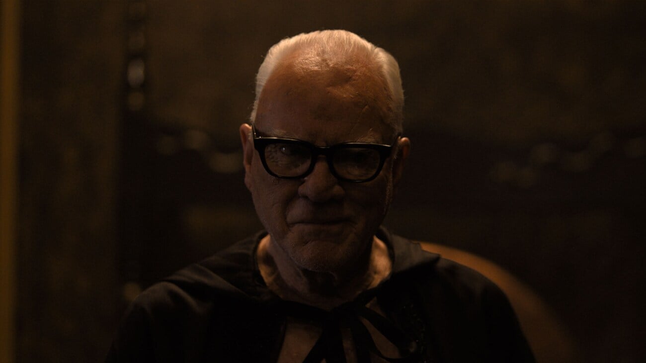An older man with white hair and glasses, wearing a dark cloak, stands in a dimly lit room with a faint smile.