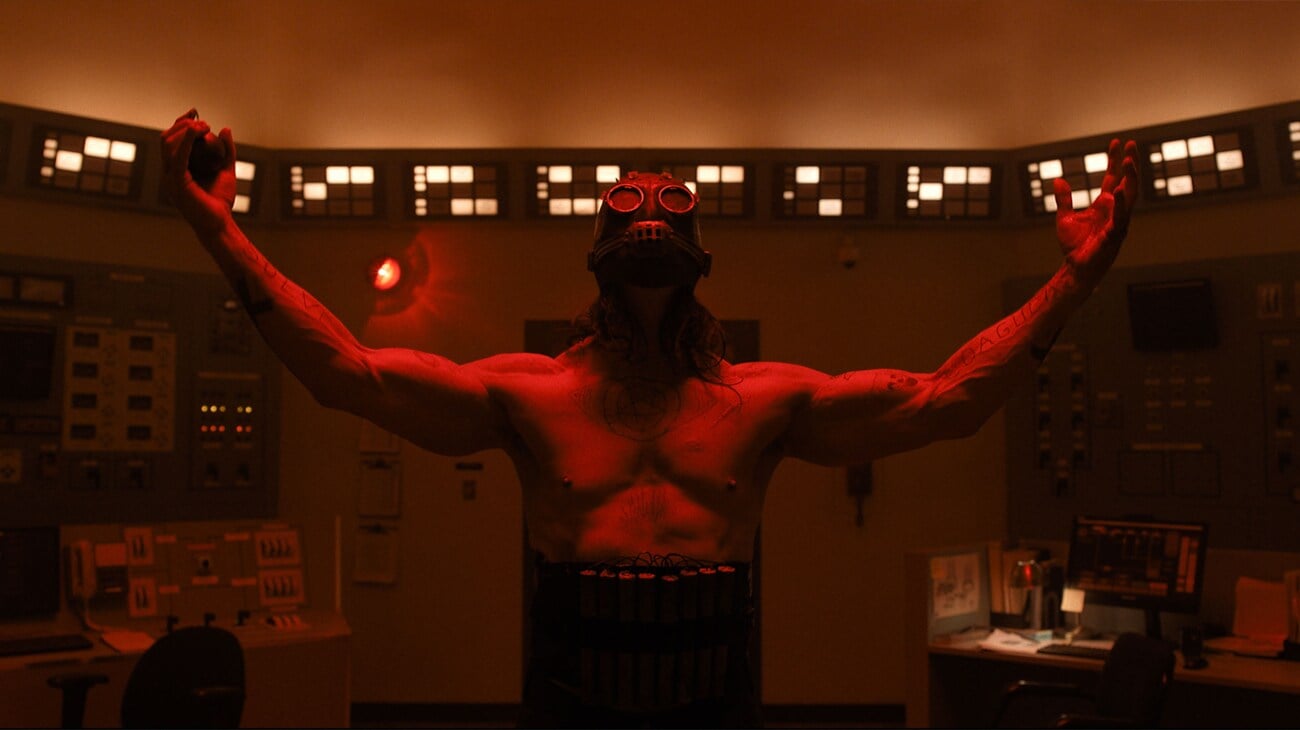 A shirtless person wearing a gas mask stands with arms outstretched in a dimly lit control room with red lighting and various control panels.