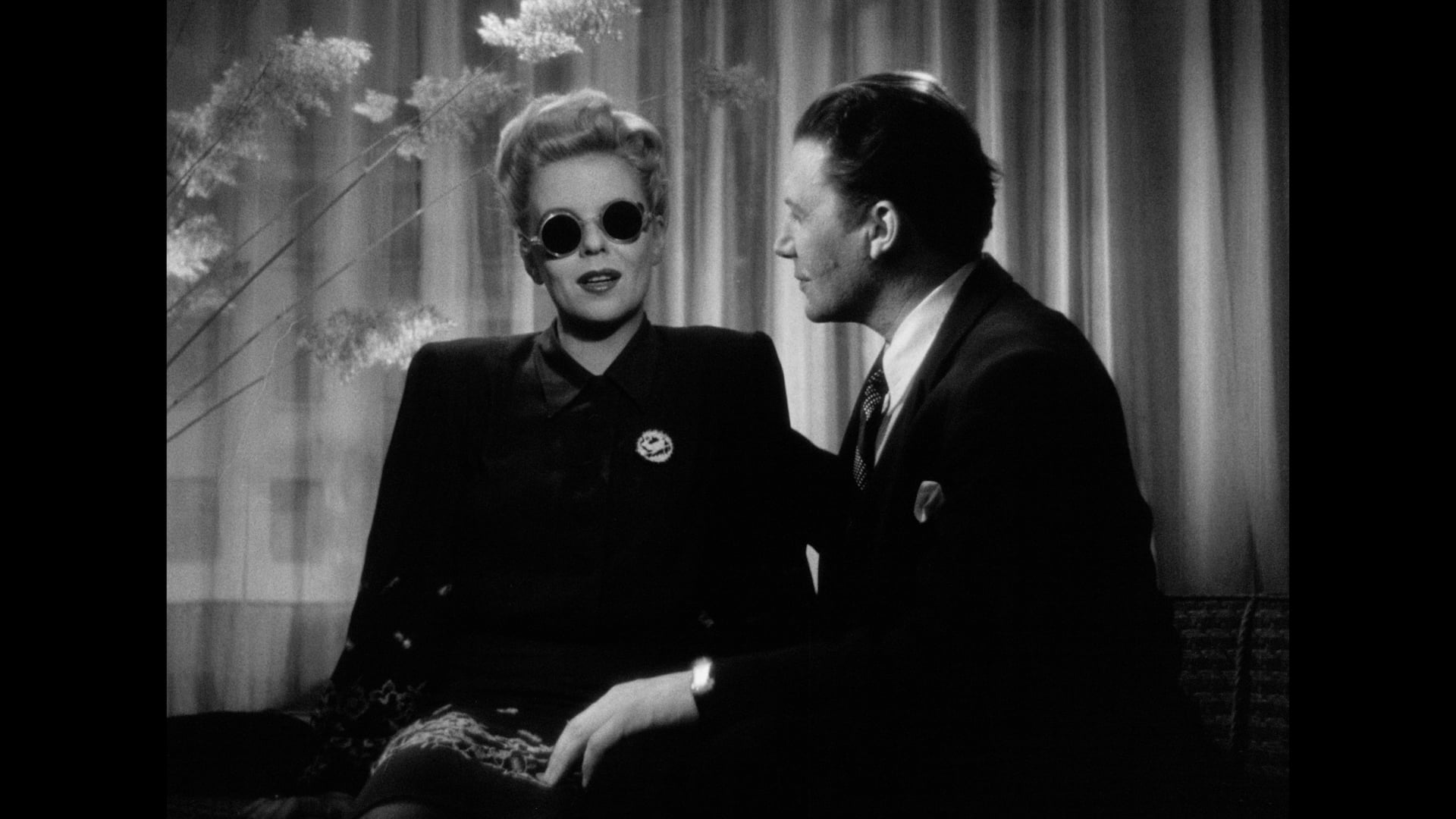 A woman wearing dark round sunglasses sits next to a man in a suit; both are indoors with sheer curtains and plants in the background.