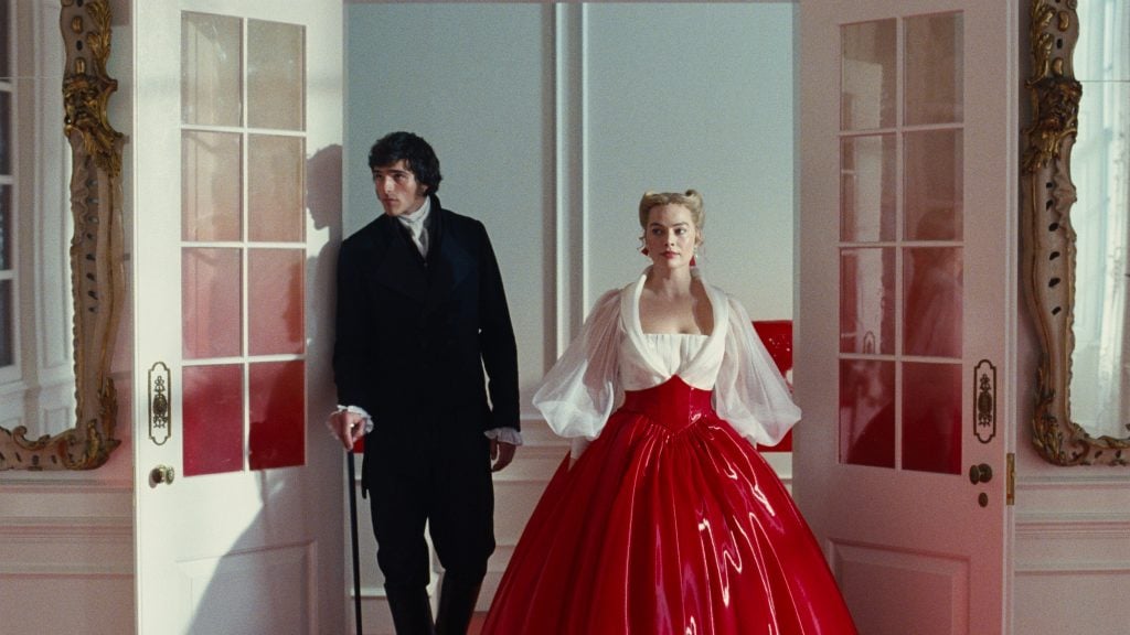 A woman in a voluminous red dress and a man in dark formal wear stand in front of white double doors in an ornately decorated room.