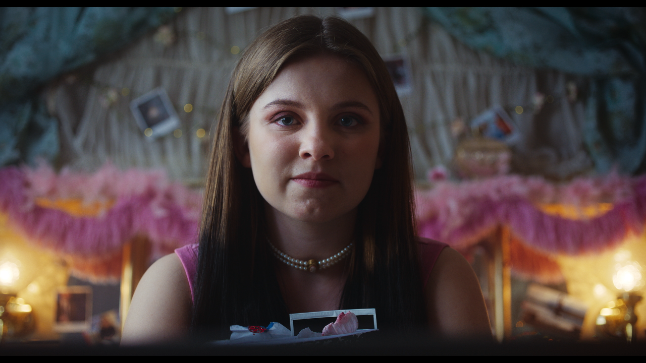 A young woman with straight brown hair and a pearl necklace sits in a softly lit room decorated with pink tulle, string lights, and hanging photos, looking directly at the camera.