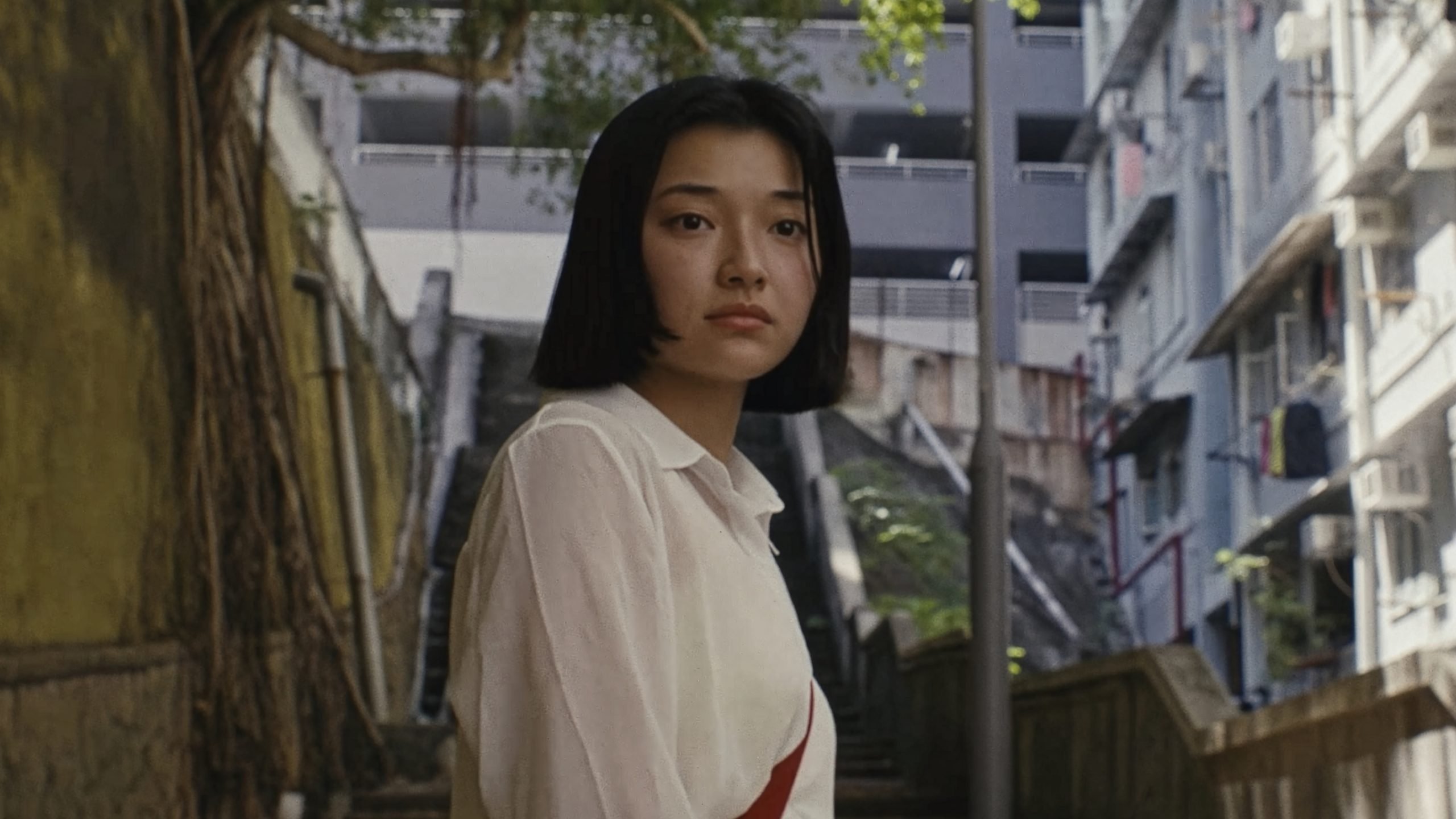 A young woman with short black hair stands outdoors near a staircase, with apartment buildings and greenery in the background.