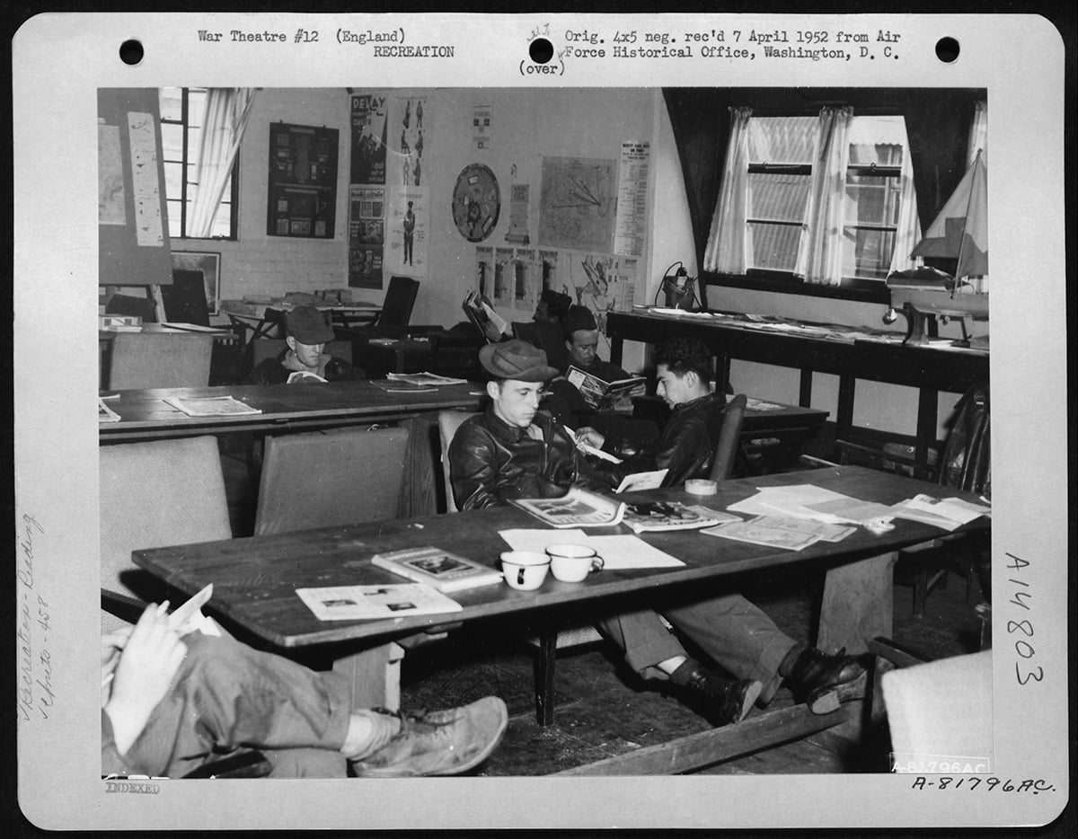 Black and white photo of servicemen relaxing in a recreation room, some reading and others seated at tables with papers and coffee cups, England, 1942.