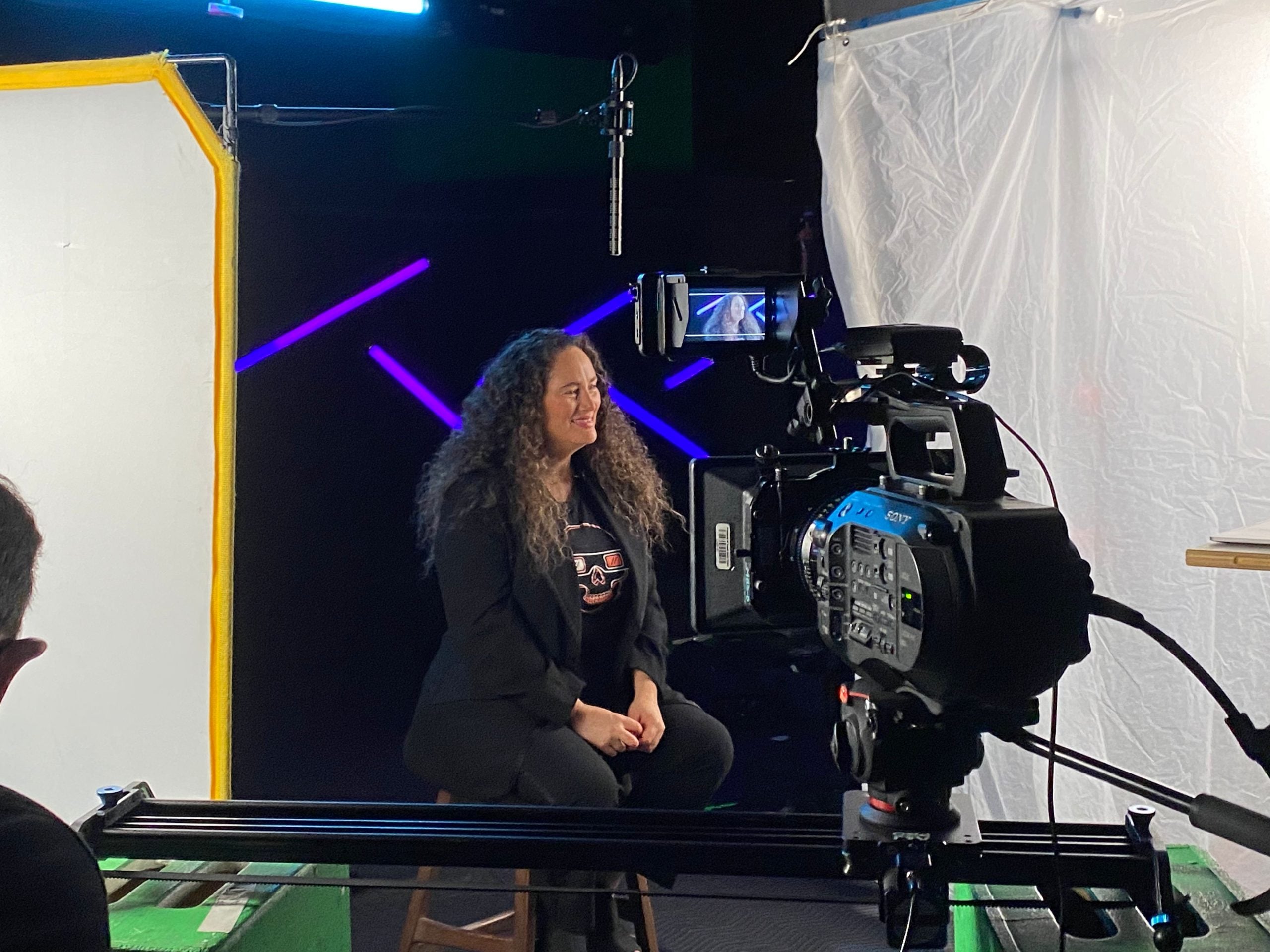 A woman with long curly hair sits on a stool in a studio, facing a professional video camera with lights and equipment set up around her.