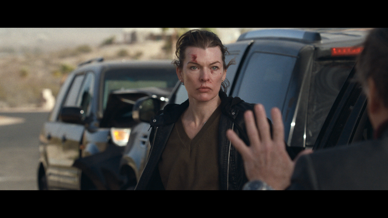 A woman with a cut on her forehead stands beside a black SUV, looking intently at a person who is raising their hand in front of her.