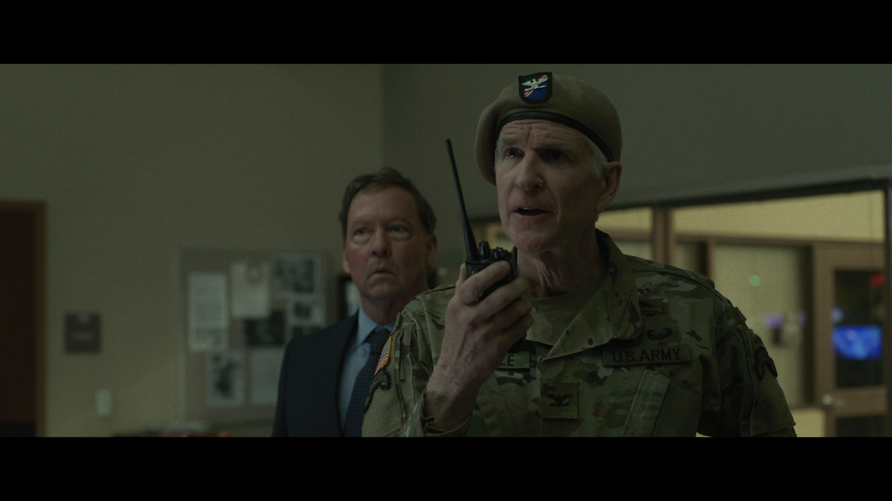 A man in a U.S. Army uniform speaks into a walkie-talkie while another man in a suit stands behind him in an office setting.