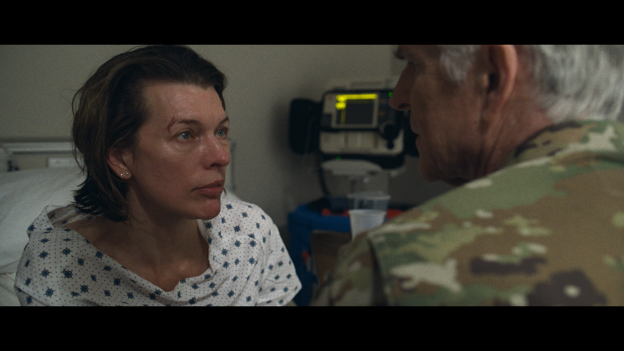 A woman in a hospital gown sits on a bed looking at a man in military uniform; medical equipment is visible in the background.