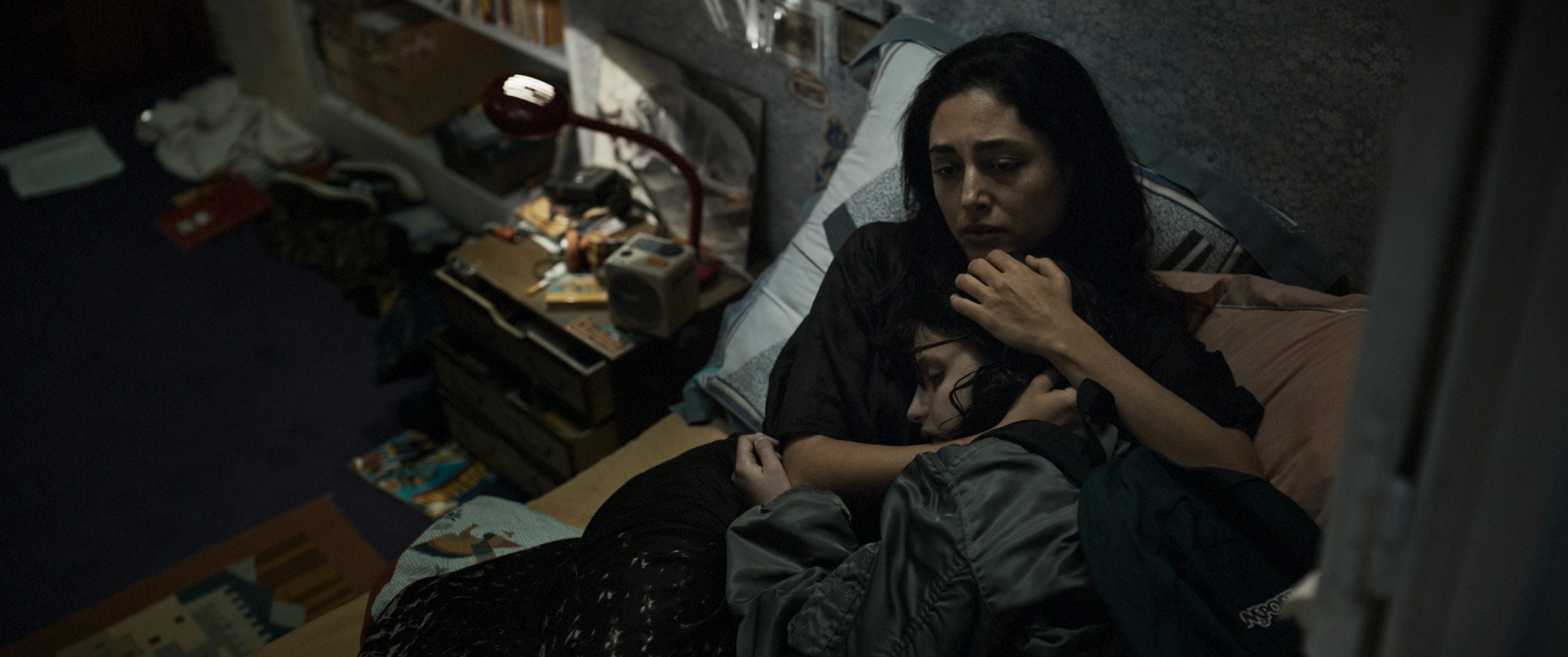 A woman sits on a bed, holding and comforting a child who is resting against her, in a dimly lit and cluttered bedroom.