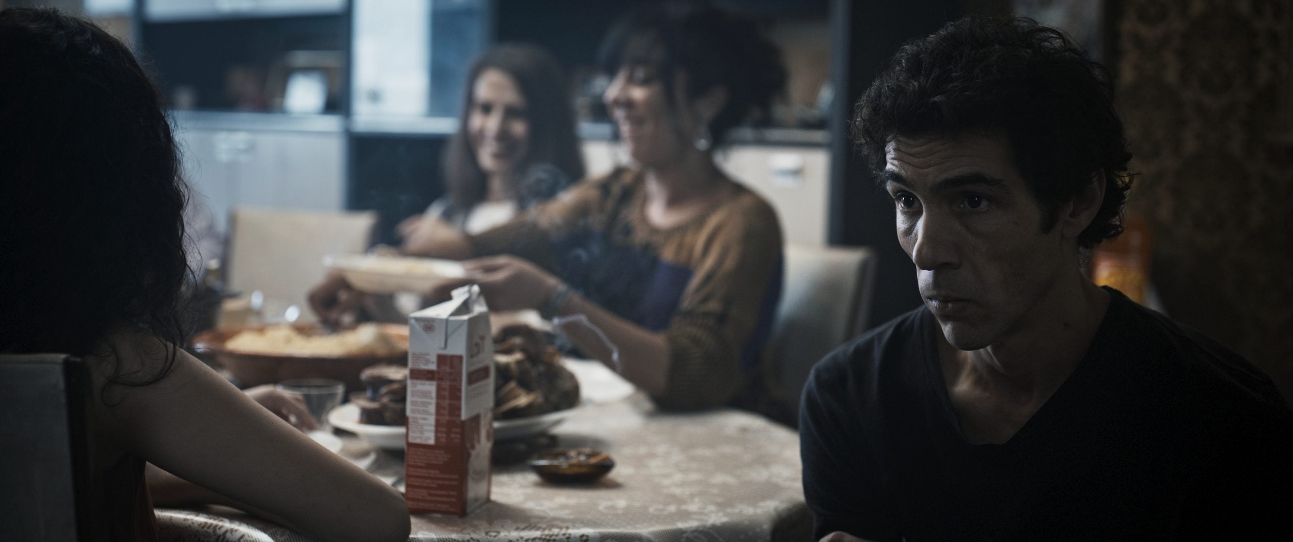 A man sits at a dining table with three women in the background, where food and drinks are laid out in a kitchen setting.