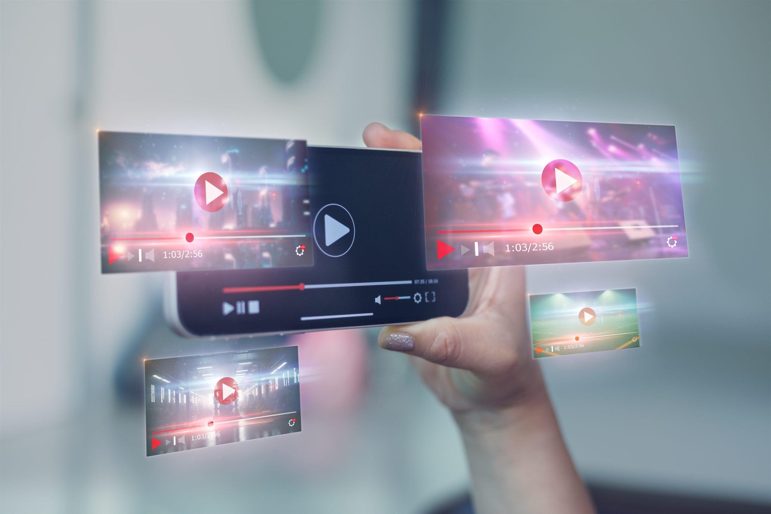 A hand holds a smartphone displaying a video player, with several digital video screens and play icons floating in the air around it, representing media streaming.
