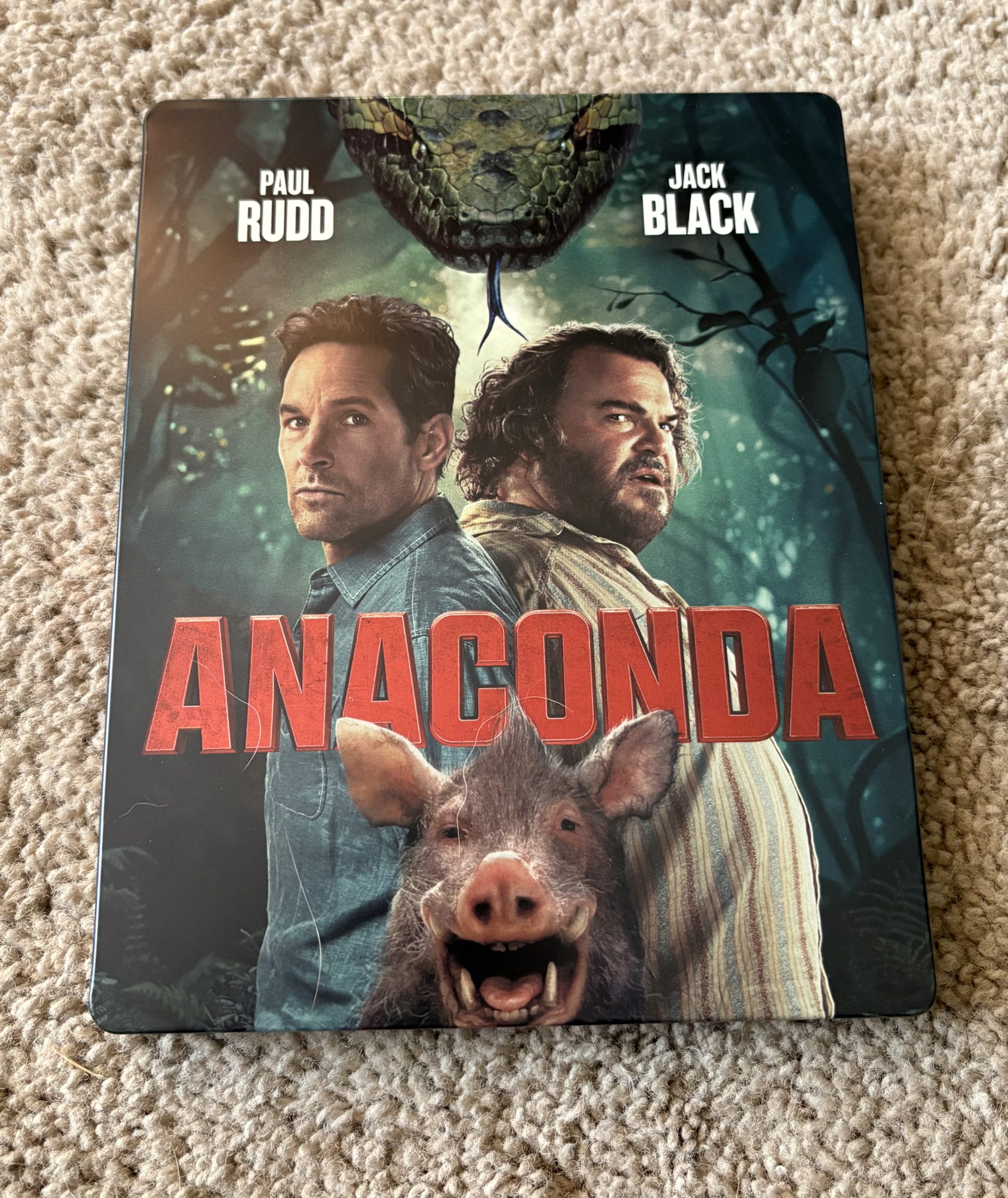 A movie case on beige carpet features Paul Rudd and Jack Black with "ANACONDA" in red text, an anaconda above, and a wild boar in the foreground.