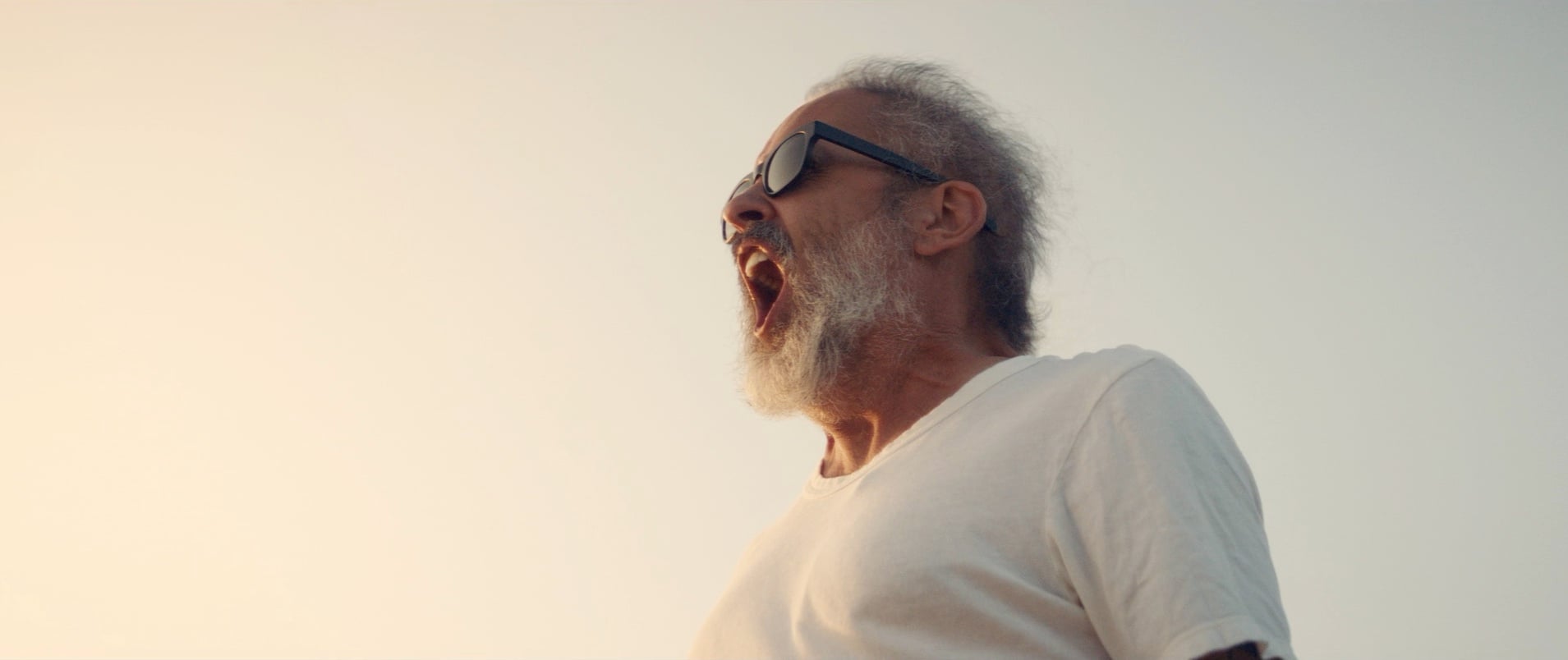 An older man with a gray beard and sunglasses, wearing a white t-shirt, stands outside with his mouth open as if shouting, against a pale sky background.