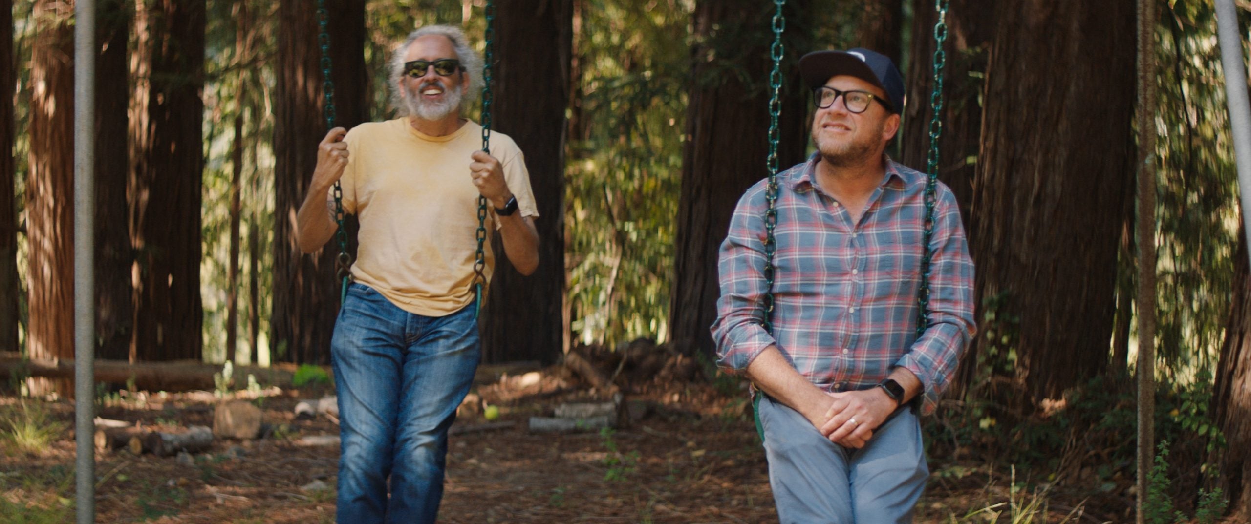 Two men sit on swings in a wooded area; one wears a yellow shirt and sunglasses, the other wears a plaid shirt, glasses, and a cap.