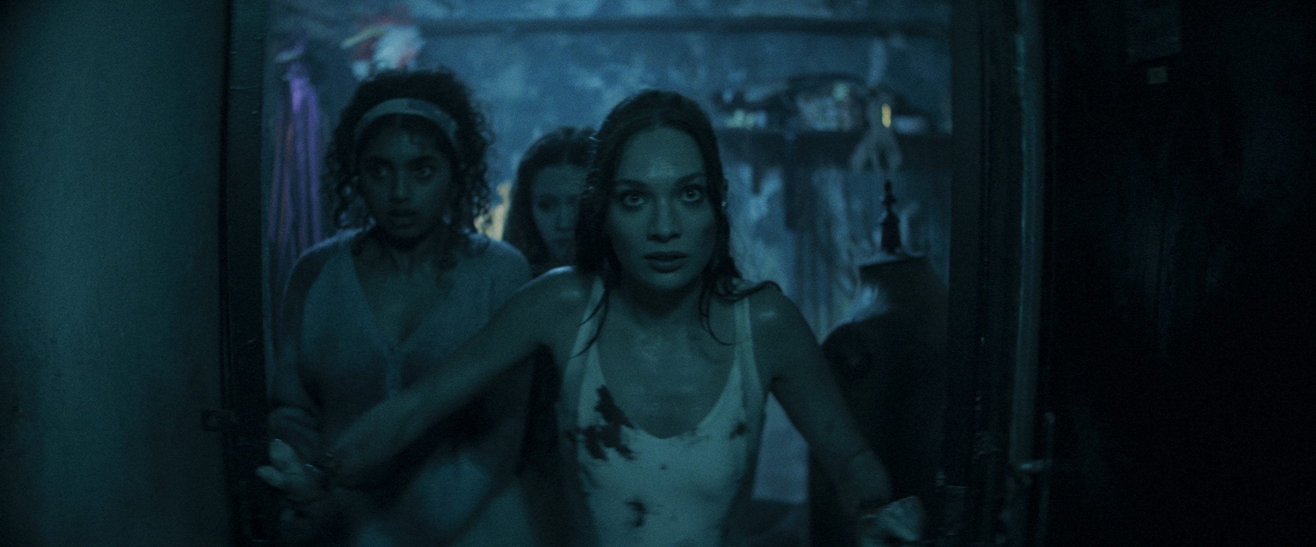 Three women with anxious expressions, covered in dirt and stains, move cautiously through a dimly lit, cluttered doorway.