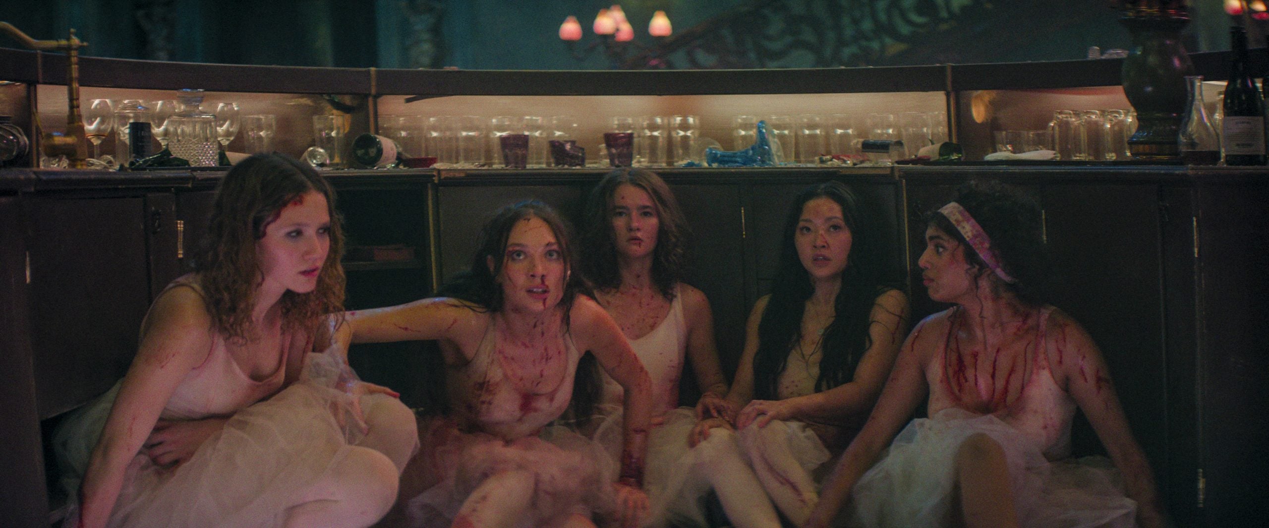 Five women in bloodstained ballet costumes sit on the floor behind a bar, looking alert and tense, with jars and bottles on shelves in the background.